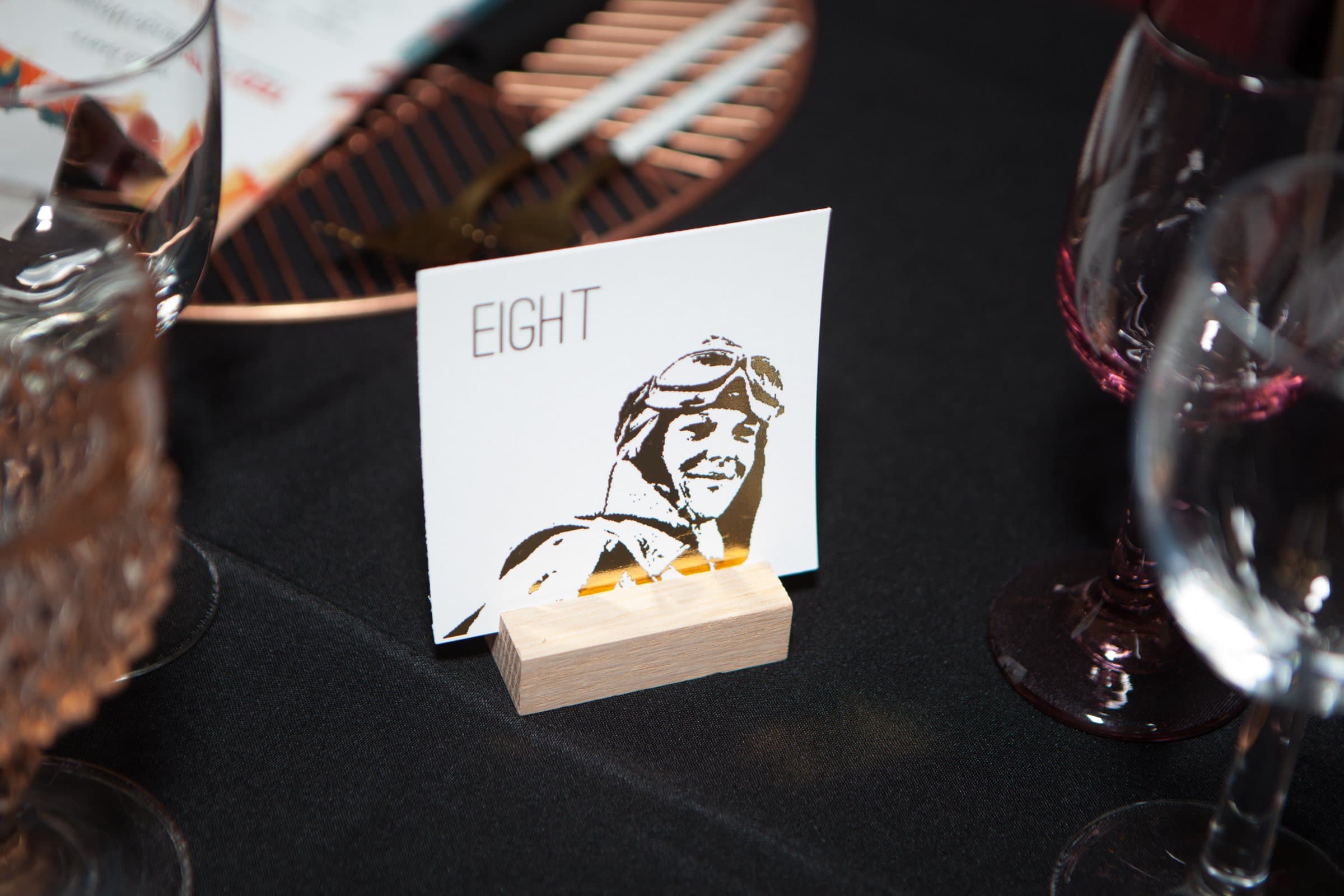 Illustrated 'Eight' table card on a dark dinner table between wine glasses